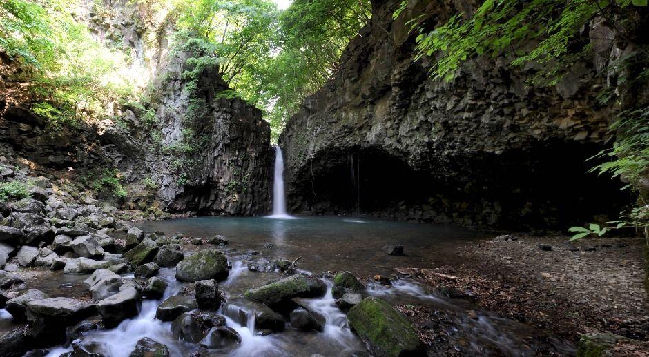 Tall waterfall cascading from basalt overhang into a calm circular pool surrounded by green forest