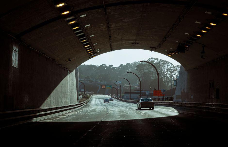 Cars driving through a long road tunnel with overhead lights leading to daylight exit