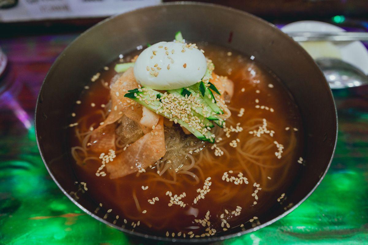 Bowl of Korean mul naengmyeon (cold broth noodles) topped with boiled egg, cucumber slices, and sesame seeds